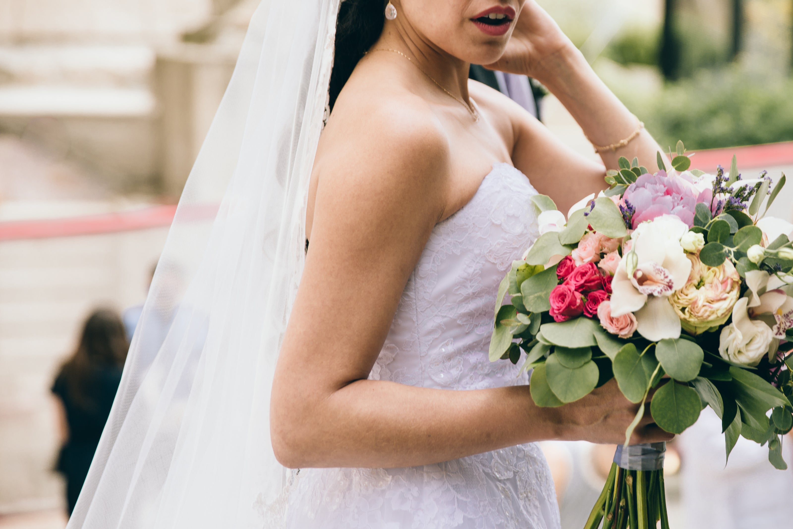 Bride wearing a white strapless wedding gown and long veil holding bridal bouquet outside of wedding venue - Orange Blossom Bridal.